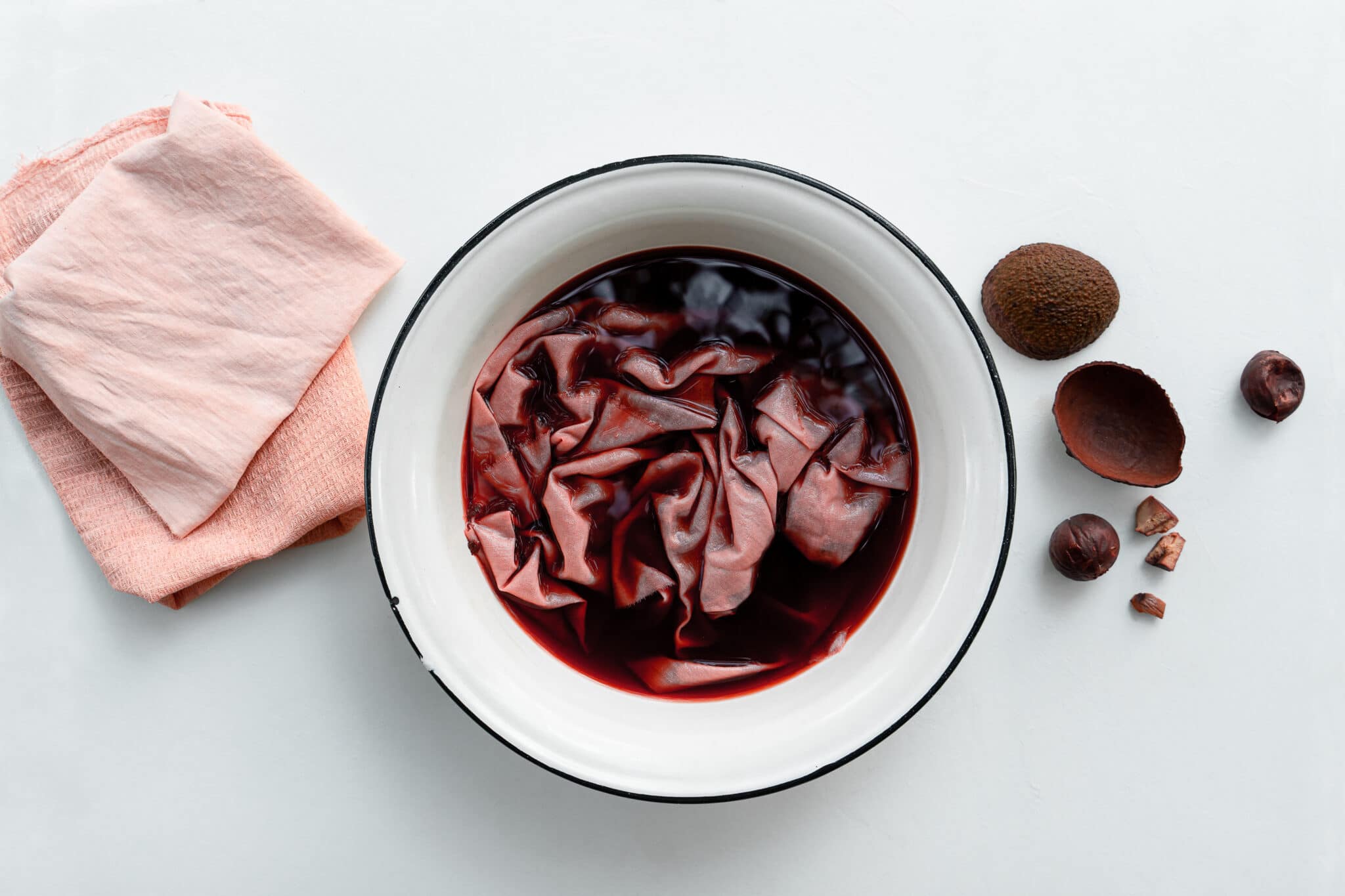 Fabric soaking in a bowl of pink natural fabric dye. Avocado skins and pits sit on the table beside the bowl.