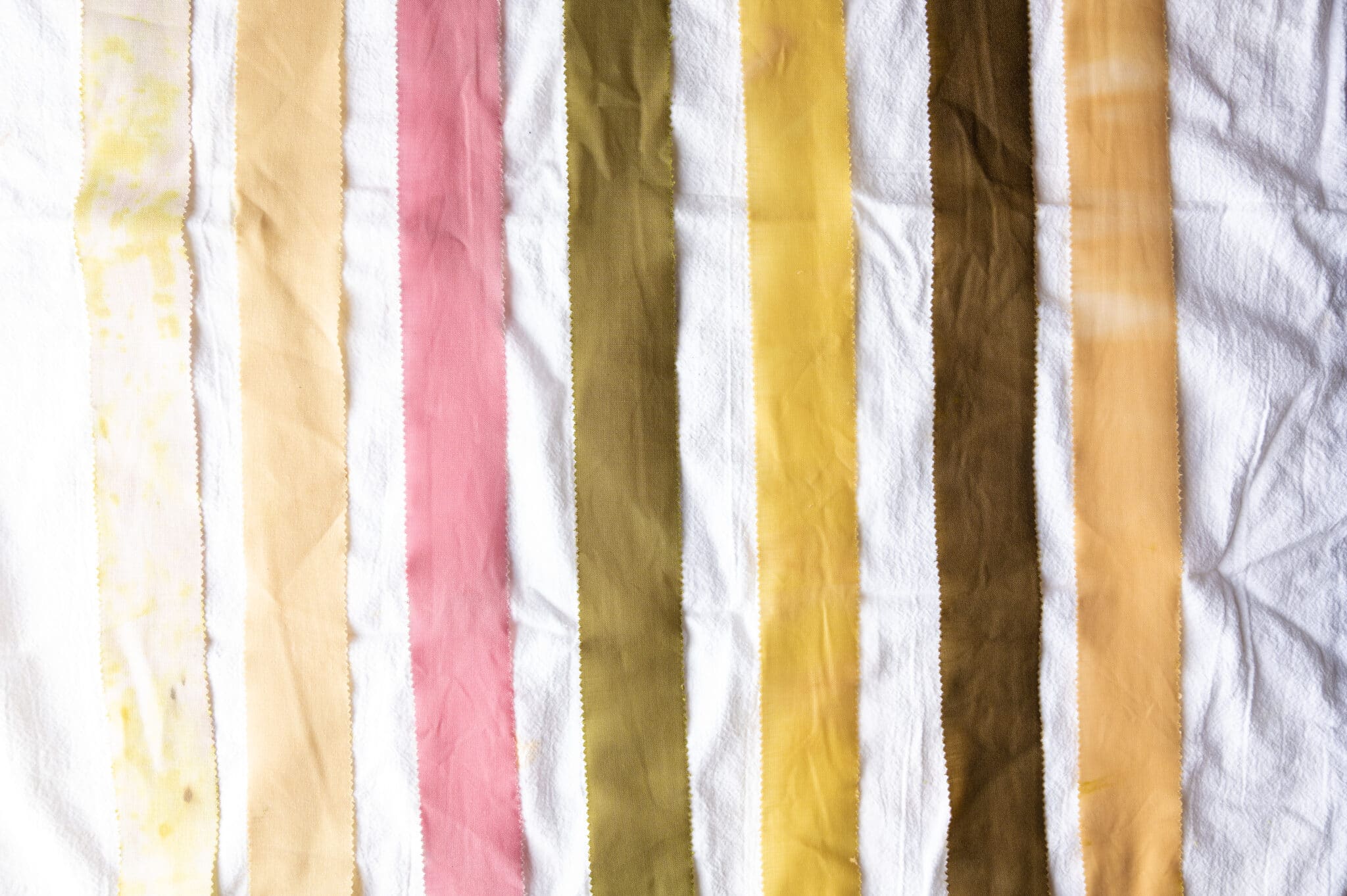 Seven strips of fabric that were dyed using natural dye made from onion skins. Each strip is a different shade, ranging from brown to yellow to pink.