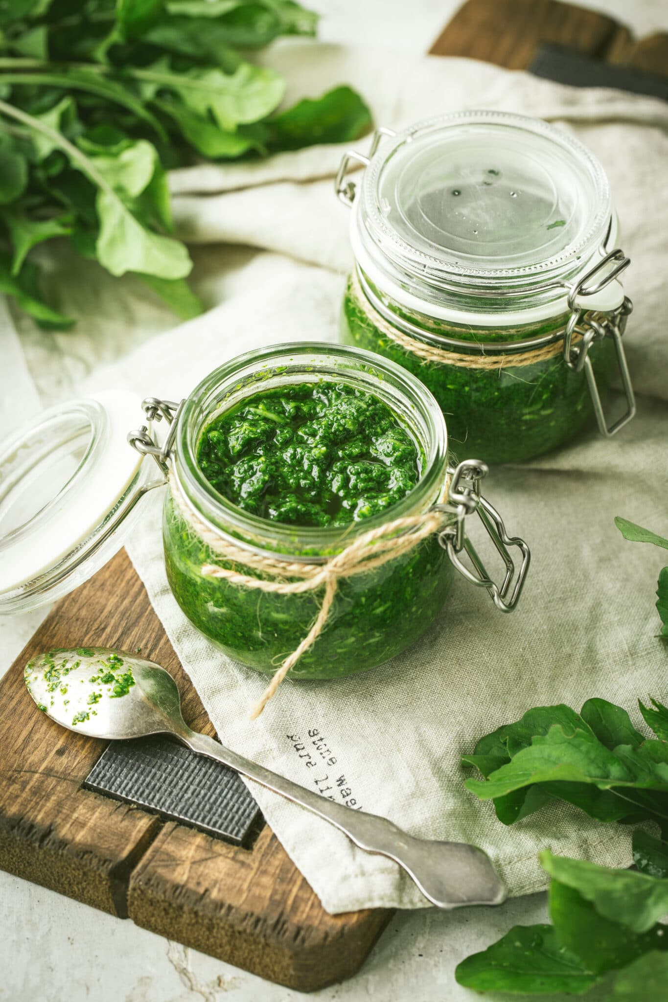 Jars of bright green homemade pest on a  rustic wooden cutting board with a linen napkin.  Fresh basil can be seen in the background.