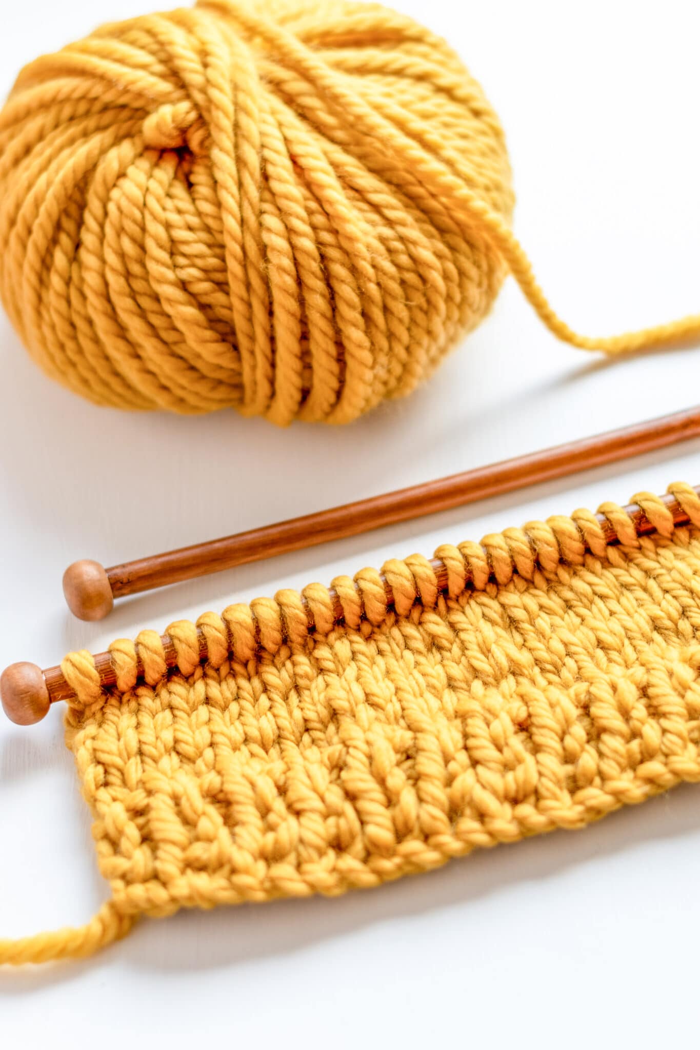 A bright yellow ball of chunky wool being knit on a wooden knitting needle to show what knitting looks like vs crochet.