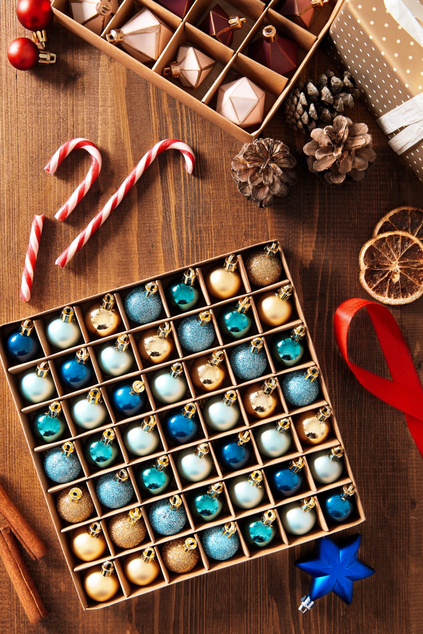 A box of gold and blue ornaments organized in a box with dividers.  Ribbon, candy canes, and pine cones are scattered around  the box. 