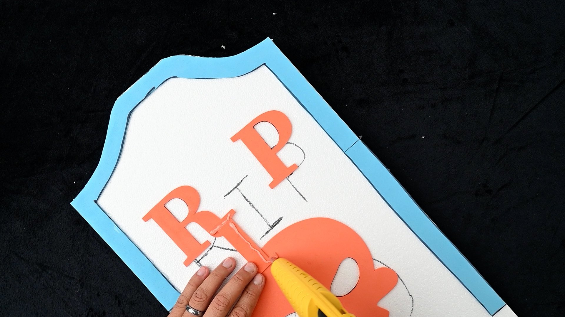 Using a hot glue gun to glue the letters for the Halloween gravestone to the styrofoam sheet.