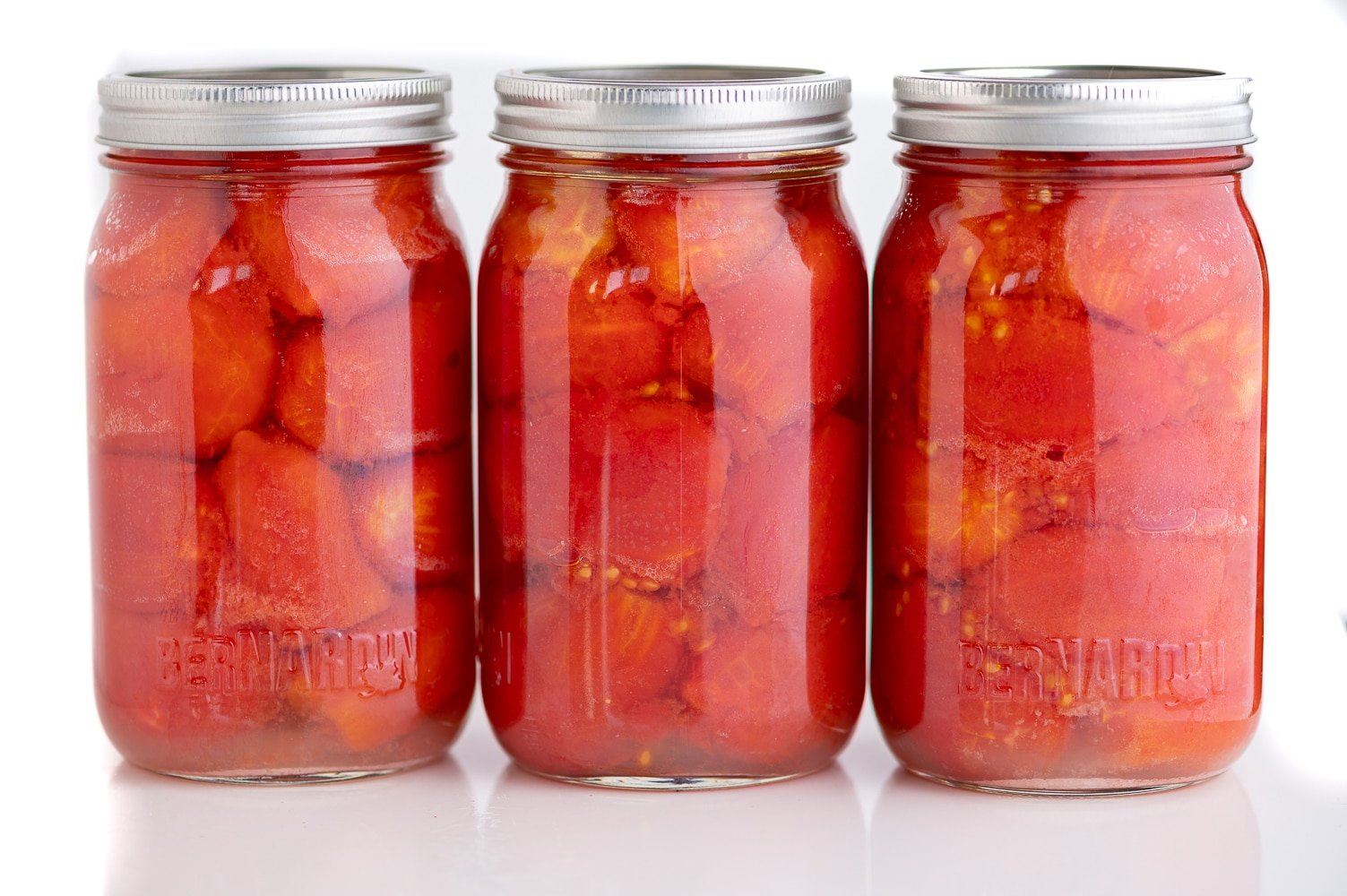three quart sized jars filled with whole tomatoes against a bright white background.