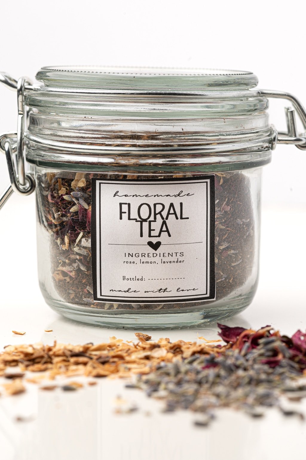 A jar of floral teas with a scattering of tea ingredients across a white table.